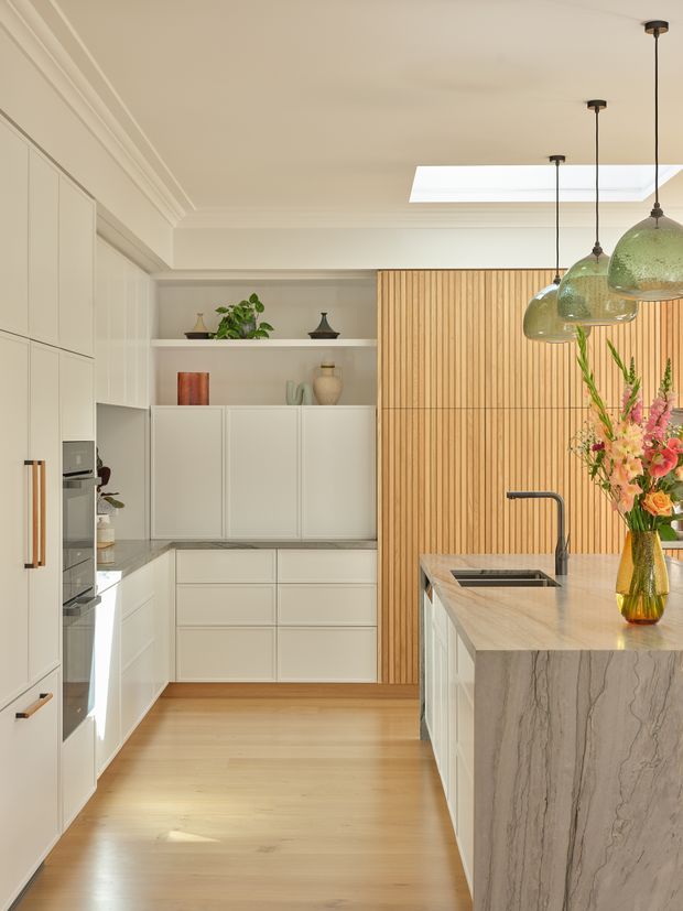 Bright, refined contemporary kitchen with a central island, pale built-in cabinetry, a veined stone island benchtop, light timber flooring, and a vertical timber feature wall. Green glass pendant lights hang above the island, while a skylight provides additional natural light. Integrated appliances and open shelving complete the clean, warm design.