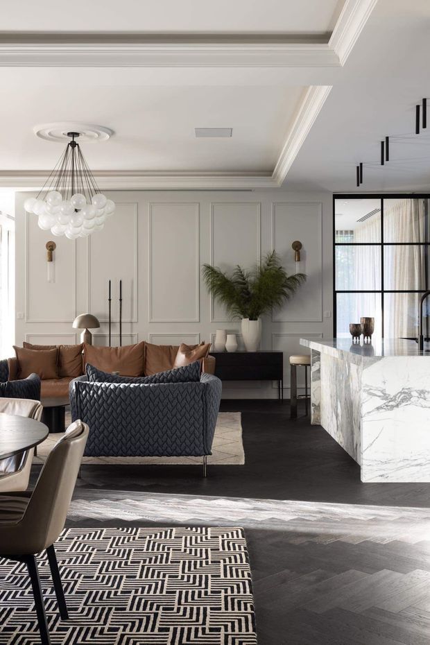 A refined open-plan living and dining interior with a sophisticated neutral palette. The room features white decorative wall panelling, ornate ceiling cornices and recessed detailing, dark herringbone timber flooring, a large marble-clad kitchen island, a tan sofa, dark textured lounge chairs, a patterned area rug, and a sculptural clustered pendant light. Black-framed internal glazing separates the living area from a brighter adjoining space.