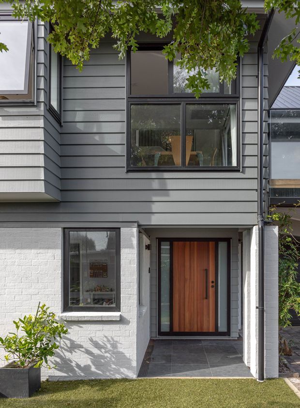 Front exterior view of a contemporary multi-level home, featuring dark grey horizontal cladding, white painted brickwork, black-framed glazing, a projecting upper window, and a warm timber pivot-style entry door set within a recessed tiled porch. Mature greenery partially frames the facade.