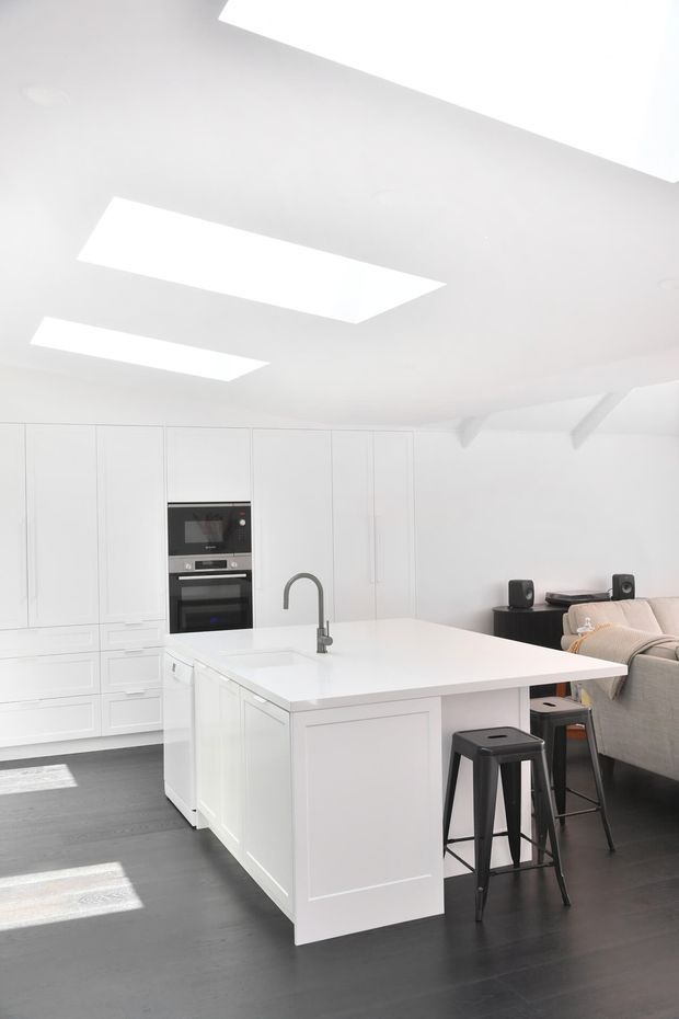 Bright open-plan kitchen with a large white island, integrated white cabinetry, built-in black oven, and a dark arched faucet. Black stools sit at the island, while a grey sofa and compact media area are visible behind it. The room has dark timber flooring, white walls and ceiling, recessed skylights or roof windows, and exposed white ceiling beams.