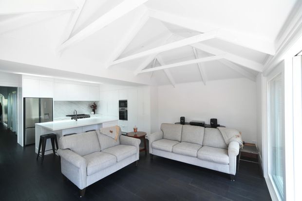 Bright open-plan interior combining a contemporary white kitchen and living room. The space features exposed white pitched ceiling trusses, dark timber flooring, pale upholstered sofas, white cabinetry, a large island with stools, stainless-steel appliances, and abundant glazing.