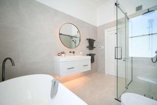 Bright contemporary bathroom featuring a freestanding white bathtub, floating double vanity with integrated drawers, large round mirror, pale stone-look wall and floor finishes, and a frameless glass walk-in shower with matte black fixtures.