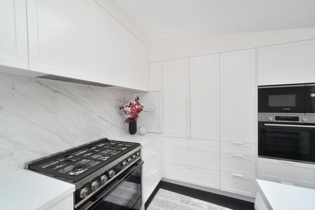 Bright, contemporary white kitchen with extensive integrated cabinetry, a large freestanding stainless-steel range cooker, built-in microwave and oven, and a full-height white marble-look splashback. The room has a clean, refined appearance with subtle black and grey accents and a pale rug visible at the floor.