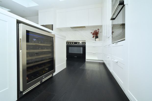 Bright, refined white kitchen with extensive built-in cabinetry, a large freestanding range, integrated appliances, marble-look splashback and benchtops, and a dark timber floor. A glass-front wine refrigerator is prominent in the foreground, with recessed ceiling lighting and a restrained monochromatic palette.