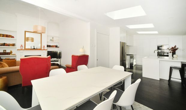 Bright open-plan kitchen, dining and living area with a large white dining table, white sculptural chairs, white cabinetry and island, dark timber flooring, built-in shelving, a red armchair and red dining chair, and multiple skylights. The space has a clean contemporary design with strong white-and-red contrast.