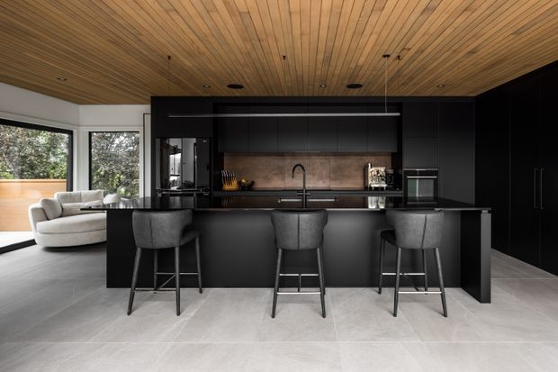 A contemporary open-plan kitchen with a large black island and three upholstered bar stools. Matte dark cabinetry, integrated appliances, a stainless-steel sink and tap, and a warm timber-lined ceiling contrast with pale large-format floor tiles. A living area and extensive glazing are visible to the left.