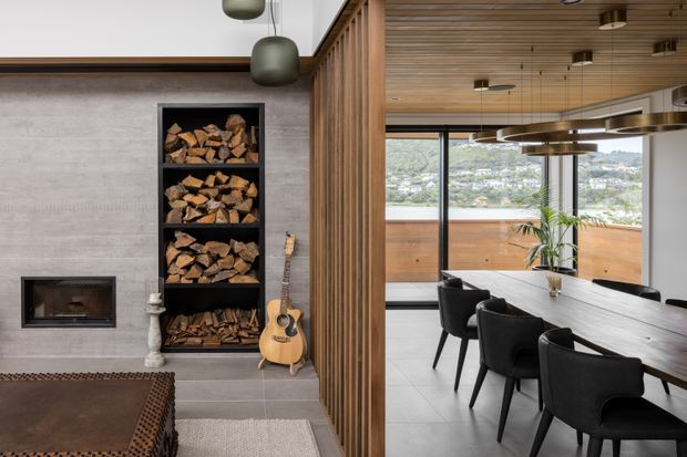 Interior view of a contemporary open-plan living and dining space. The living area features a concrete fireplace wall with built-in firewood storage, timber detailing, and a guitar, while the adjoining dining area has a long dark table, black upholstered chairs, pendant lighting, and extensive glazing overlooking a green hillside.
