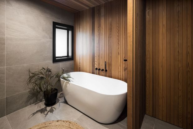 A refined modern bathroom featuring a freestanding white oval bathtub, large neutral stone-look wall and floor tiles, warm vertical timber wall and ceiling panelling, a black-framed window, and a potted plant. The space has a warm, spa-like atmosphere with concealed or minimal fixtures and strong timber detailing.