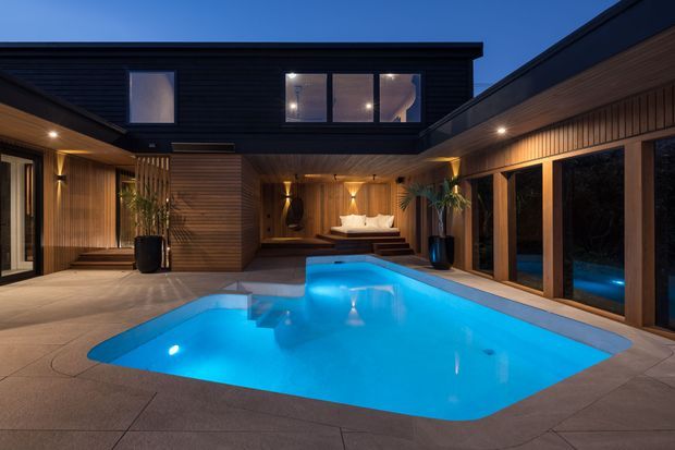 Twilight view of a contemporary courtyard residence centered around a brightly illuminated swimming pool. Dark horizontal cladding, warm timber-lined soffits and walls, extensive glazing, recessed lighting, and an integrated covered outdoor lounge create a refined indoor-outdoor setting.