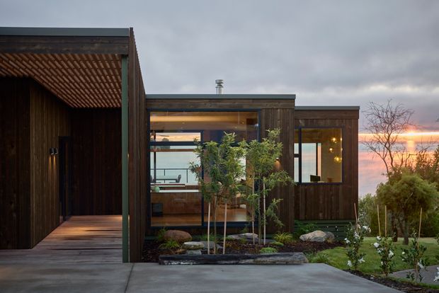 A compact modern single-storey home with dark timber cladding and large glazed openings, arranged around a sheltered entry porch with a slatted timber pergola. The low, rectilinear form is set within a landscaped garden, with exposed interior spaces visible through the glazing and a cloudy sunset sky beyond.