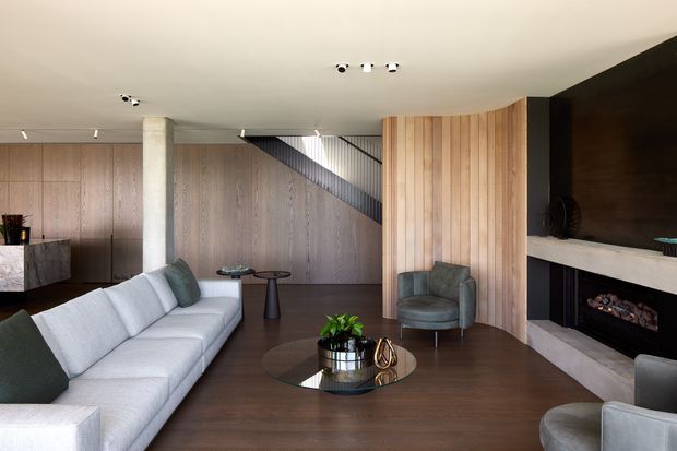 A refined contemporary living room with a large pale sectional sofa, dark timber flooring, full-height timber wall panelling, a curved vertical timber screen, and a dramatic black fireplace wall with a pale stone mantel. A stairway is partially visible behind the timber panelling, while a round glass coffee table and sculptural armchairs complete the room.