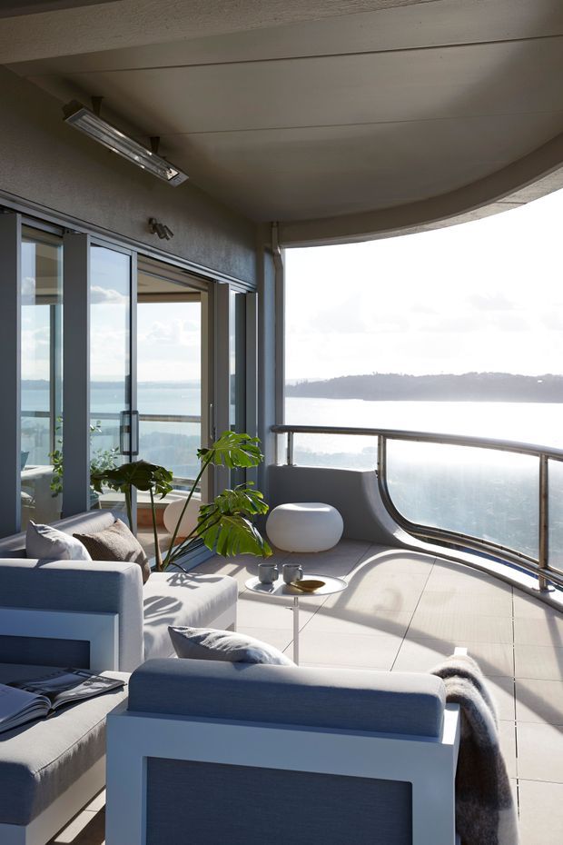 A bright coastal living room or enclosed balcony overlooking a body of water. The space features large sliding glass doors, curved floor-to-ceiling glazing, a rounded balcony, pale tiled flooring, white and blue upholstered lounge chairs, a small round coffee table, and potted tropical foliage. A low wooded shoreline is visible beyond the glazing.