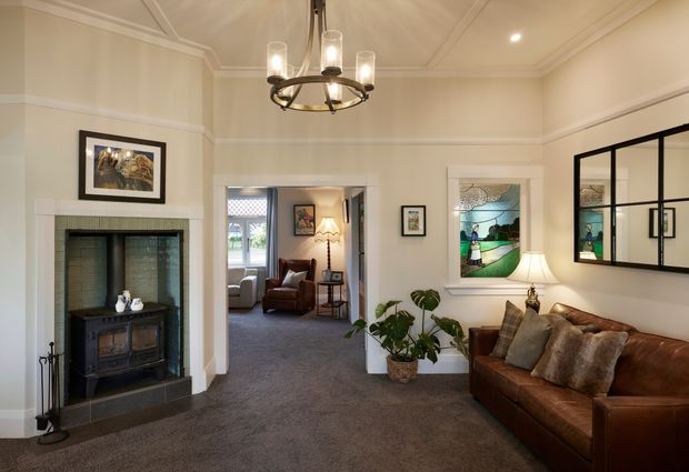 Elegant traditional living room with cream-painted walls, decorative wall mouldings, a tiled fireplace surround with black freestanding stove, dark carpet, and a brown leather sofa. A glazed internal doorway opens to a secondary sitting area, while stained-glass artwork, framed art, plants, and a central chandelier add character.