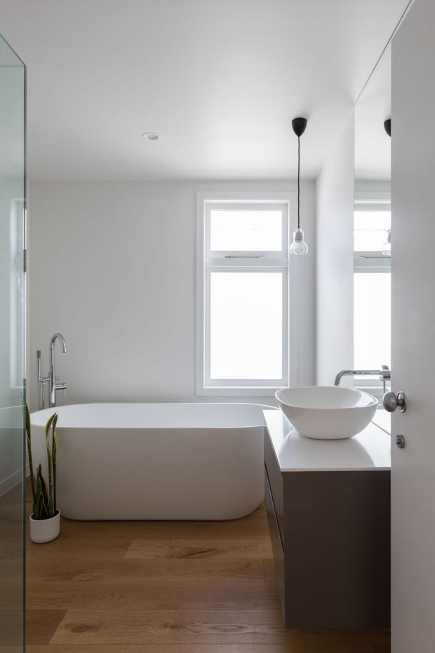 A bright, minimalist bathroom viewed through an open doorway, featuring a freestanding white bathtub, a white vessel basin on a dark vanity, chrome fixtures, pale walls, large mirrored surfaces, timber-look flooring, and simple pendant lighting. A narrow window provides natural light, while a small potted plant adds a restrained natural accent.