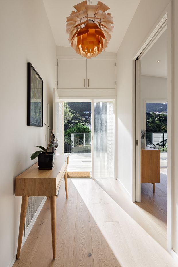 Bright, narrow residential hallway or entry passage with pale timber flooring, white walls, a slim timber console, framed artwork, and a sculptural warm-toned pendant light. Full-height glazed doors open toward an exterior deck and landscape, while a mirrored or reflective opening on the right reveals part of an adjacent room. Built-in white overhead cabinetry provides storage above the exterior door.