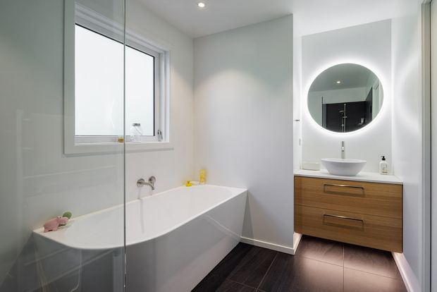 Bright contemporary bathroom featuring a freestanding white bathtub beside a frameless glass shower screen, a wall-mounted timber vanity with white vessel basin, and a large illuminated round mirror. The room has pale walls, recessed ceiling lighting, dark timber-look floor tiles, and a clean minimalist fit-out.