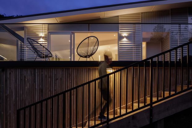 Twilight exterior view of a contemporary, multi-level residence with a low-pitched roof, expansive glazed openings, horizontal cladding and warm interior lighting. A timber privacy screen and black metal balustrade frame an elevated walkway or deck, with occasional outdoor seating visible through the glazing.
