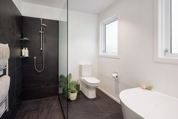 Bright contemporary bathroom with a frameless glass walk-in shower, dark large-format tiled shower wall and floor, white toilet, freestanding white bathtub, and white-framed windows. A small plant and neatly arranged towels add softness to the otherwise high-contrast scheme.