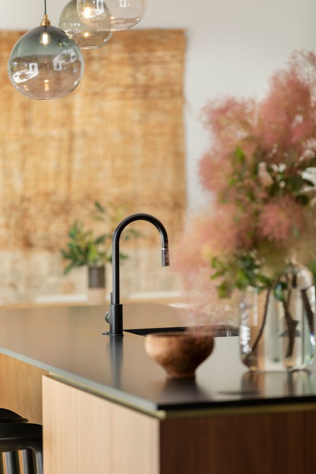 Close-up view of a refined kitchen island featuring a tall matte-black curved mixer tap, dark stone benchtop, and light timber cabinetry. Warm woven timber-toned wall cladding forms the backdrop, with glass pendant lights overhead and soft pink floral foliage adding decorative colour. The image has a warm, contemporary feel with a focus on the tap and island joinery.