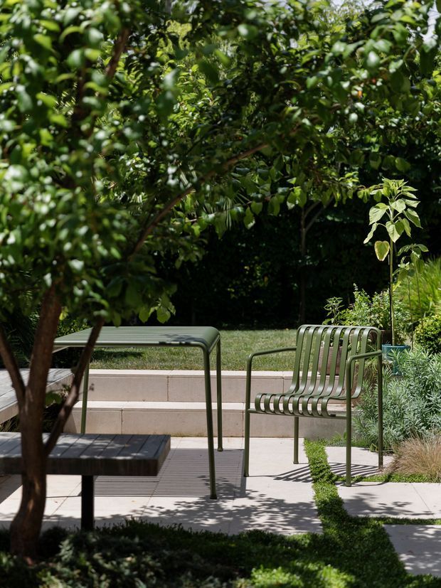 A landscaped outdoor courtyard or garden seating area with a contemporary metal table and two matching slatted metal chairs on a paved terrace. Mature leafy planting provides shade, with raised concrete garden beds, clipped hedging, ornamental grasses and shrubs creating a private, refined garden setting.