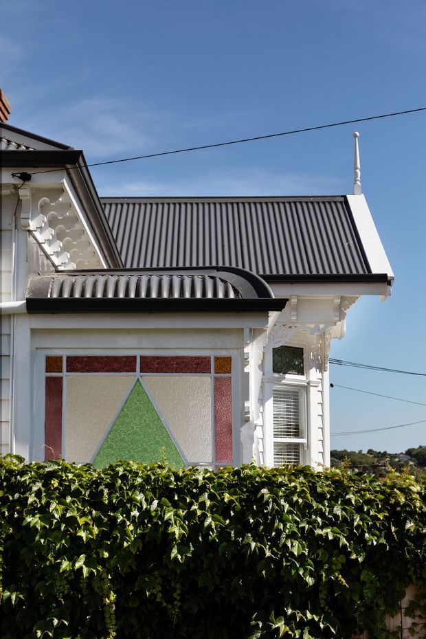 Exterior close-up of an ornate historic house, featuring a steep corrugated metal gable roof, white decorative timber trim and brackets, a bay-style window, and a prominent geometric stained-glass panel. A dense hedge partially screens the lower facade.