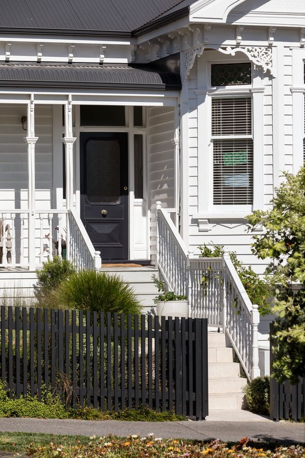 Sunlit close-up of the front entrance of a heritage-style timber-clad house. The facade features white horizontal weatherboards, ornate decorative brackets and trim, a dark corrugated roof, a black glazed entry door, Victorian-style porch columns and balustrades, and a raised stair approached through a dark vertical-slat front fence. Established planting borders the entry.