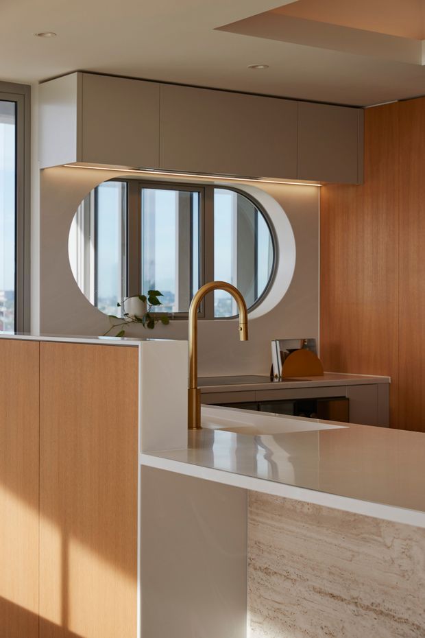 A refined contemporary kitchen viewed across a large island. The space features warm timber cabinetry, taupe and cream flat-panel joinery, a pale stone benchtop with a textured stone waterfall end, an integrated curved brass tap, concealed under-cabinet lighting, and a distinctive oval interior window opening onto tall glazing. Soft natural light creates a warm, polished atmosphere.