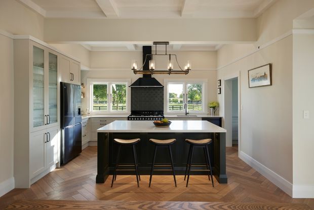 Bright, spacious kitchen with a large central island and three bar stools, white perimeter cabinetry with glass-front storage, a dark rangehood and cooker, and a stainless-steel refrigerator. The room features warm herringbone timber flooring, white walls and coffered ceiling detailing, black metal accents, pendant lighting, and garden views through the windows.