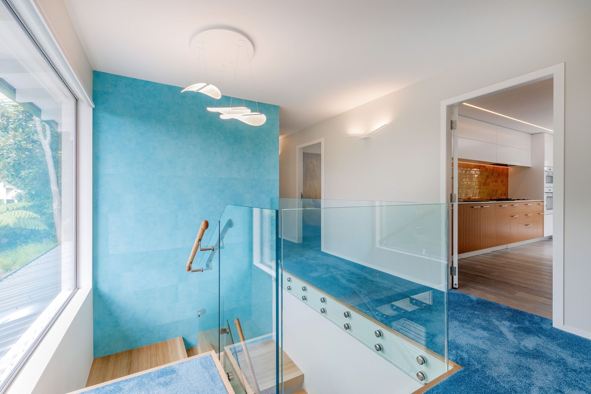The feature wallpaper is Elitis Wallpaper Natives Maori. It runs floor to ceiling over two stories. The wallpaper reflects Oceania. The Diphy_P3 Pendant by Grok looks like sea creatures swaying and floating and the wing lights in the hall evoke sea birds hovering above the ocean.