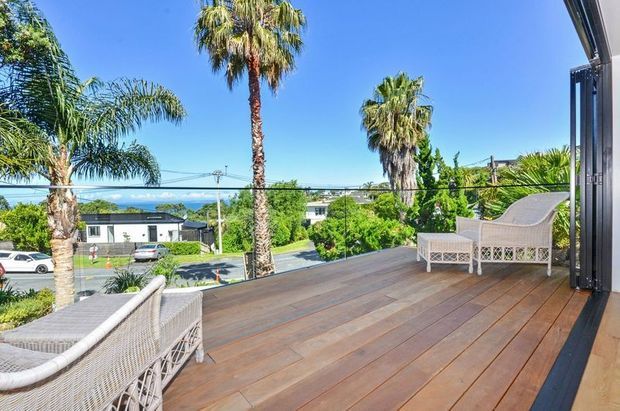 An elevated outdoor timber deck or terrace viewed from a large open glazed doorway, furnished with white woven outdoor seating. The deck overlooks a leafy suburban neighbourhood and distant blue water or coastline, with tall palm trees and a bright clear sky. The scene has a relaxed coastal outdoor-living character.