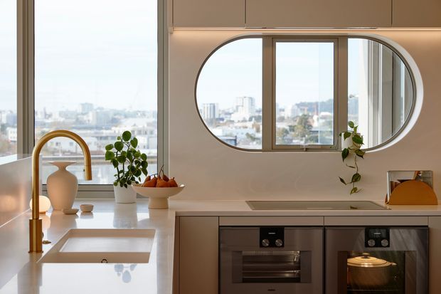 Bright contemporary kitchen with a white stone island and integrated sink, brushed brass gooseneck tap, dark lower cabinetry, built-in appliances, and a distinctive oval interior window opening onto a city view. Natural light, indoor plants, and warm under-cabinet lighting create a refined, airy atmosphere.