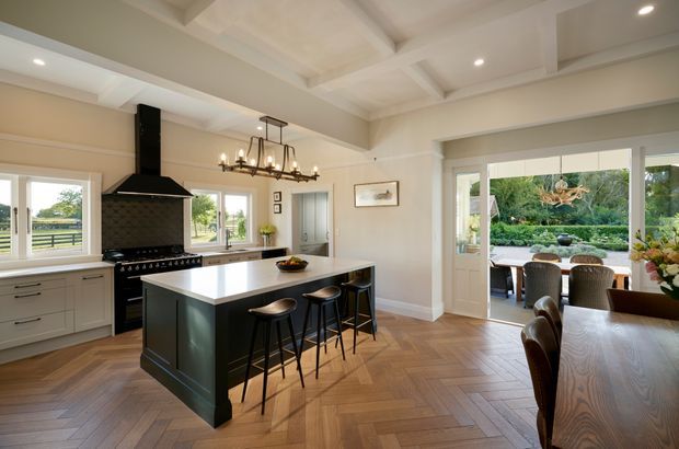 A bright, spacious open-plan kitchen and dining area with a large dark island, integrated seating, a black rangehood and cooker, pale cabinetry, and a patterned timber floor. The room features a coffered ceiling, pendant chandelier lighting, and wide glazed doors opening to an outdoor dining area and garden.