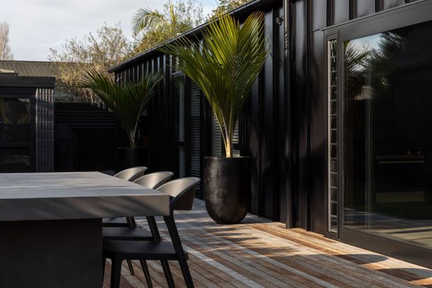 A contemporary dark-clad outdoor dining terrace beside a black-framed glazed building. A long dining table with modern chairs sits on a timber deck, while tall potted palms add greenery. Large sliding glass doors reveal part of the interior kitchen and reflect the surrounding planting. The setting has a refined, high-contrast indoor–outdoor feel.