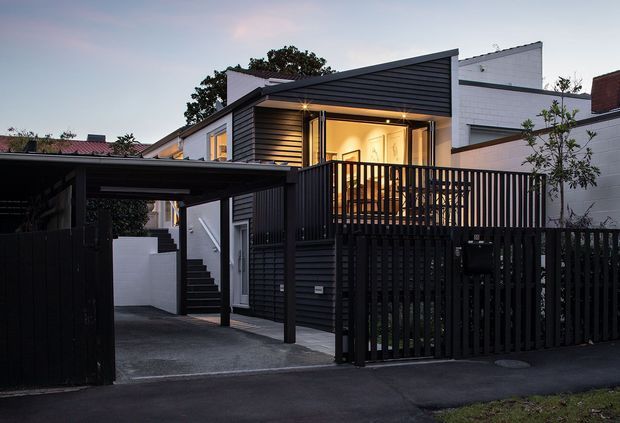 Street-facing dusk view of a compact contemporary multi-level residence with dark horizontal cladding, a sharply angled roof canopy, warm interior lighting, a raised timber deck behind a vertical-slat privacy fence, and an attached dark carport. White masonry walls and neighbouring structures are visible around the home.