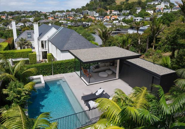 Elevated view of a contemporary residential property with a rectangular swimming pool, paved pool terrace, covered outdoor lounge, and dark-clad pavilion. Dense tropical planting surrounds the outdoor spaces, while a pitched-roof main house and a suburban hillside neighbourhood are visible beyond.