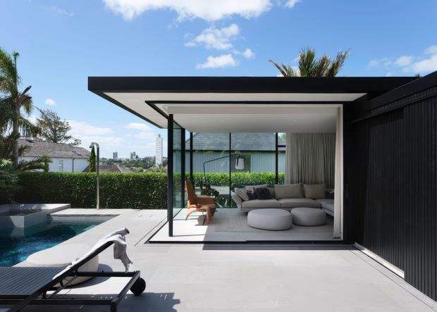 Contemporary poolside pavilion or outdoor living room with a flat roof, extensive floor-to-ceiling glazing, and a dark vertical-clad side wall. The bright, minimalist interior contains a large neutral sectional sofa and rounded ottomans, opening directly onto a pale paved terrace beside a swimming pool. Palm trees, hedges, and distant urban buildings are visible beyond the property.
