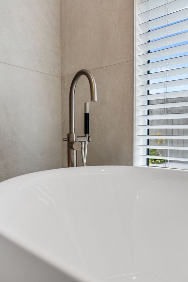Close-up of a contemporary bathroom featuring a white freestanding bathtub, a brushed metal floor- or wall-mounted tub filler with hand shower, large beige-grey stone-look wall tiles, and white horizontal plantation shutters beside the tub. The space is clean, bright, and minimally styled.