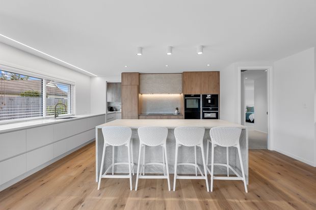 Bright contemporary kitchen with a long white island and four white bar stools, pale wood flooring, extensive white cabinetry, and a warm timber kitchen wall with integrated black appliances. Large horizontal windows, recessed ceiling lights, and a doorway to an adjacent room are visible.