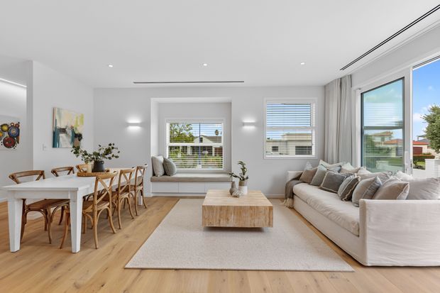 Bright open-plan living and dining area with white walls, light timber flooring, a white dining table with woven wooden chairs, a large pale sectional sofa, a textured neutral rug, and a light timber coffee table. Multiple windows with blinds and curtains provide abundant daylight, while recessed and wall-mounted lighting create a clean, contemporary interior.