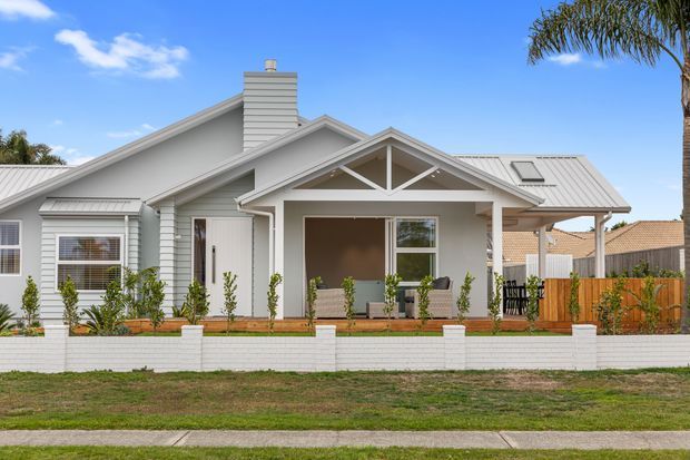 Single-storey residential house viewed from the street, featuring pale grey horizontal weatherboard cladding, white trim, a gabled roof with a prominent covered front porch, white metal roofing, and a low white brick garden wall. The front yard includes simple raised planting beds and a timber privacy fence. Bright blue sky and a palm tree are visible in the background.