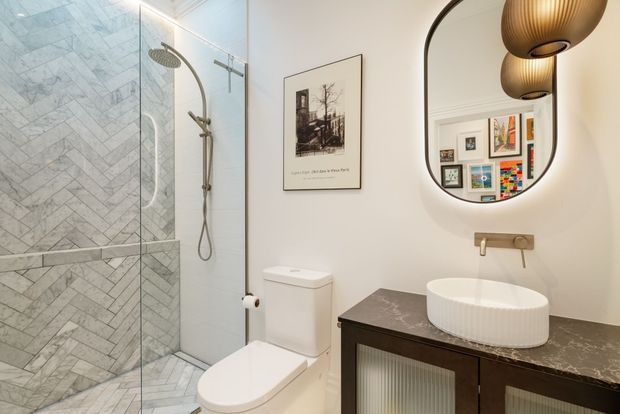 A refined contemporary bathroom featuring a walk-in glass shower with grey marble-look herringbone wall and floor tiling, a white toilet, and a dark timber vanity with a dark stone benchtop. The vanity has a white vessel basin and wall-mounted brushed brass tapware. An arched mirror, framed artwork, and sculptural brass pendant lights add decorative detail.