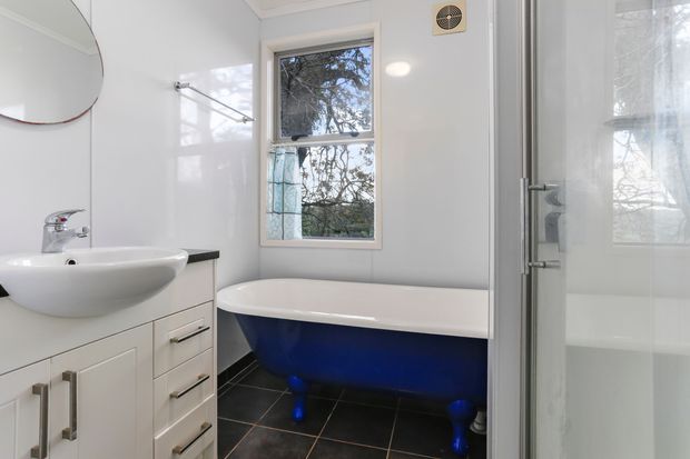 Bright compact bathroom with a white vanity and vessel basin, freestanding clawfoot bathtub with a deep blue exterior, dark tiled floor, white wall finishes, a central window, and a glass shower enclosure. The room has a clean traditional character with a bold blue feature bathtub.
