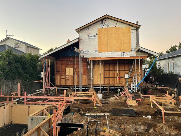 A residential timber-framed house undergoing major construction or renovation. The building is raised above an excavated site with exposed timber framing, plywood sheathing, Tyvek weatherproofing, temporary supports, and unfinished foundations. Neighbouring houses are visible on both sides.