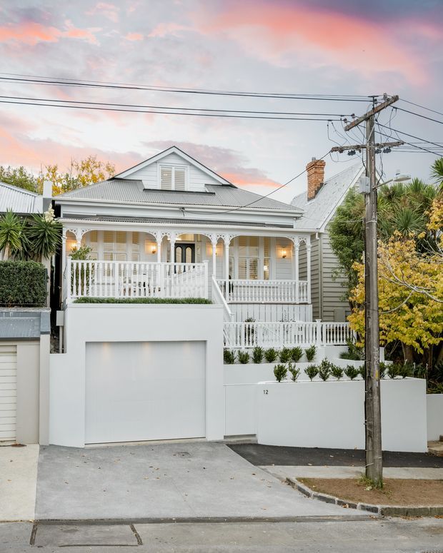 Street-facing exterior view of a renovated Victorian-era cottage or bungalow with a white-painted timber facade, corrugated metal gabled roof, decorative veranda, ornate fretwork, balustrades, upper dormer, brick chimney, and an integrated street-level garage. The home sits in an established suburban streetscape with overhead utility wires and mature planting.