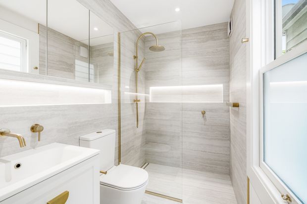 Bright contemporary bathroom featuring a white vanity and bathtub, wall-hung toilet, frameless glass shower enclosure, large pale grey stone-look wall and floor tiles, mirrored cabinetry, and brushed brass/gold tapware and shower fittings. White-framed windows and integrated horizontal lighting contribute to an airy, refined appearance.