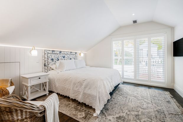 Bright, softly styled bedroom with a vaulted sloping ceiling, white bedding, a floral upholstered headboard, built-in white cabinetry, bedside table, woven armchair, layered area rugs, and large white plantation-shuttered windows. The room has a calm coastal-traditional feel with warm timber flooring and neutral finishes.