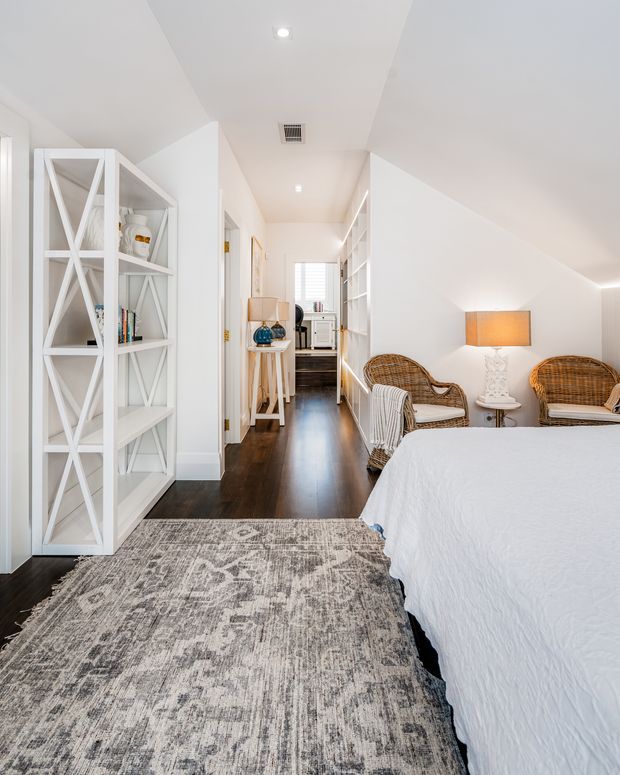 Bright, compact bedroom viewed from the foot of the bed, with a white upholstered or dressed bed, dark timber flooring, a large muted grey patterned rug, white built-in shelving and cabinetry, and wicker lounge chairs beside a small side table and warm table lamp. The room has a sloping ceiling and a narrow corridor leading toward a kitchenette or work area, creating an airy, coastal-inspired retreat.
