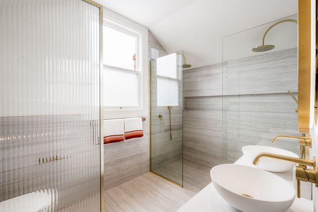Bright contemporary bathroom with a walk-in glass shower, two white vessel basins, brushed gold fixtures, and large grey wood-grain stone-look wall and floor finishes. A fluted translucent glass partition separates the vanity area from the shower, while a window provides natural light.