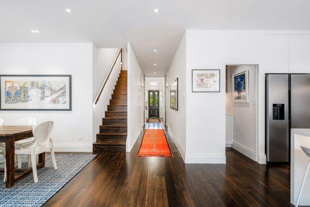 Bright open-plan interior showing a dining area, central hallway, staircase, and partial kitchen. The space has white walls and ceiling, dark timber flooring, a timber stair with slim metal balustrade, recessed downlights, framed artwork, and a vivid red-orange runner rug leading toward a glazed entry door.