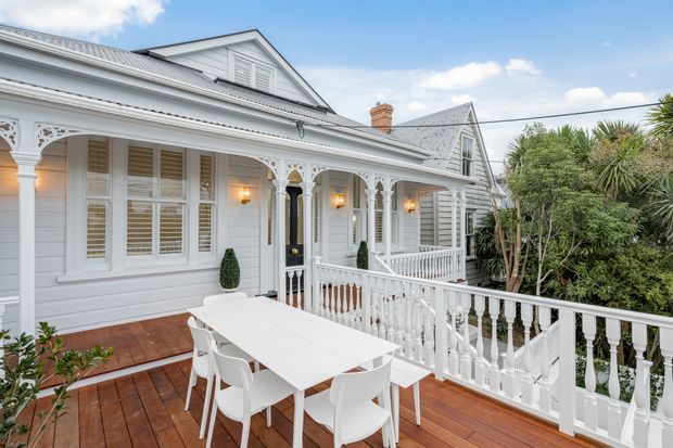 A light-filled exterior view of a heritage-style timber house with an elevated timber deck and dining area. The facade features white horizontal weatherboards, ornate decorative verandah brackets, white balustrades, shuttered windows, a dark entry door, and pitched gabled roofs. Lush mature planting surrounds the deck, with neighbouring rooflines visible in the background.